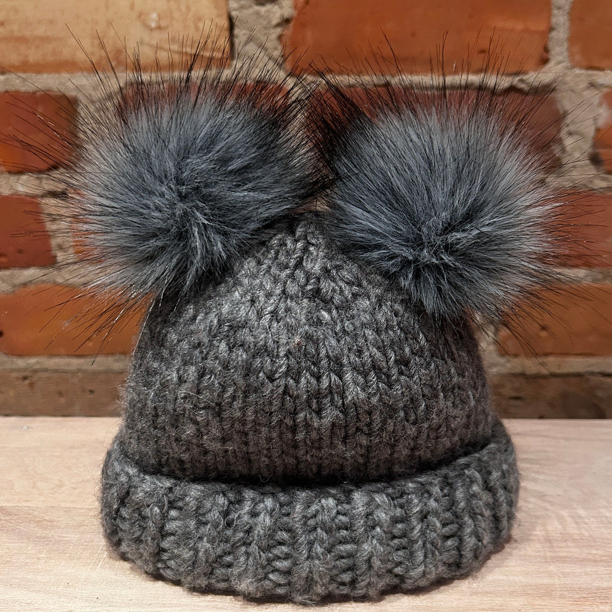 A small dark gray knit beanie hat with two small fluffy gray faux-fur pom-poms on top, displayed on a light wood surface with a red brick wall background.