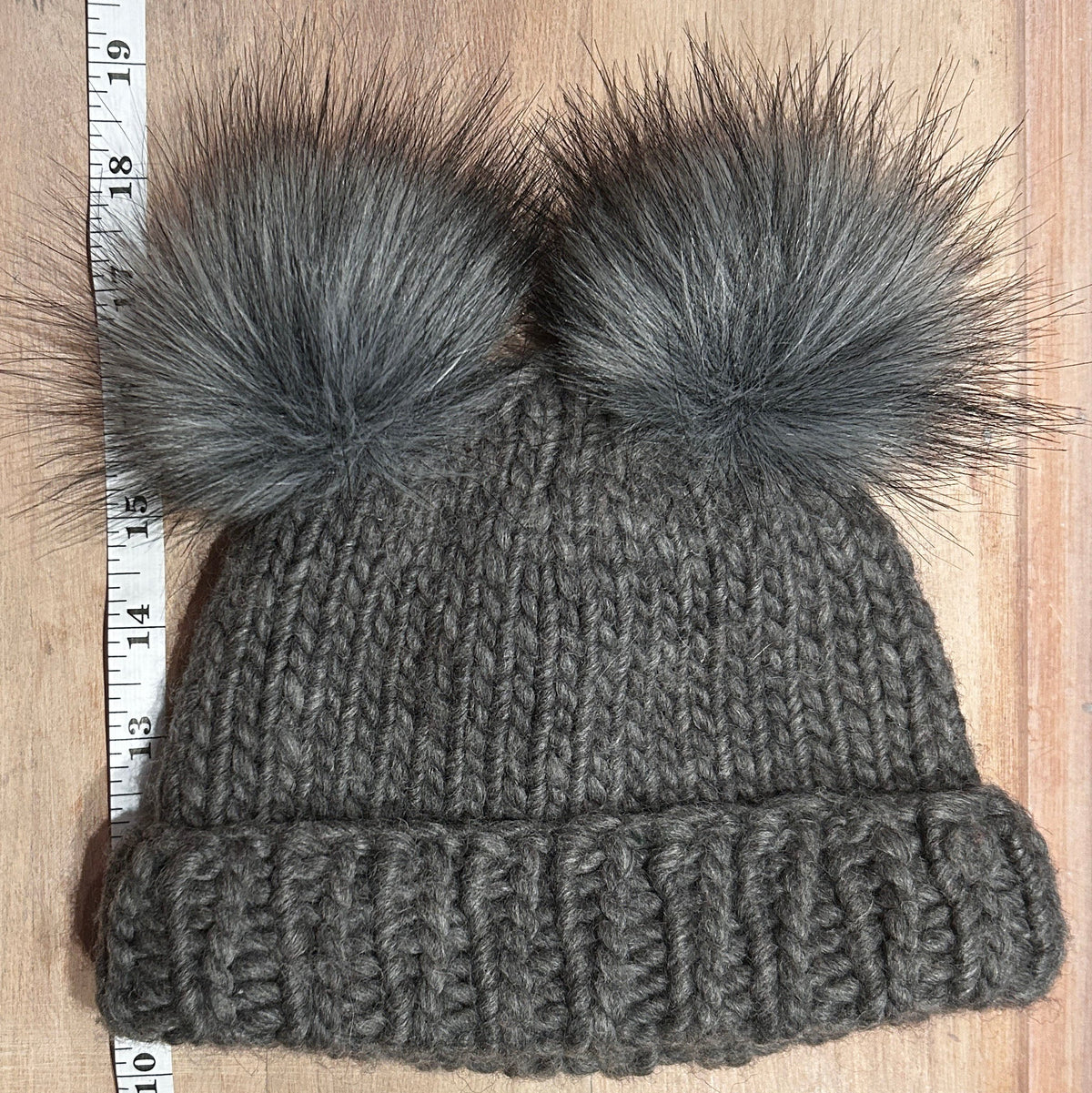 A small gray knit beanie with two small gray faux-fur pom-poms, laid flat on a light wood surface next to a measuring ruler.