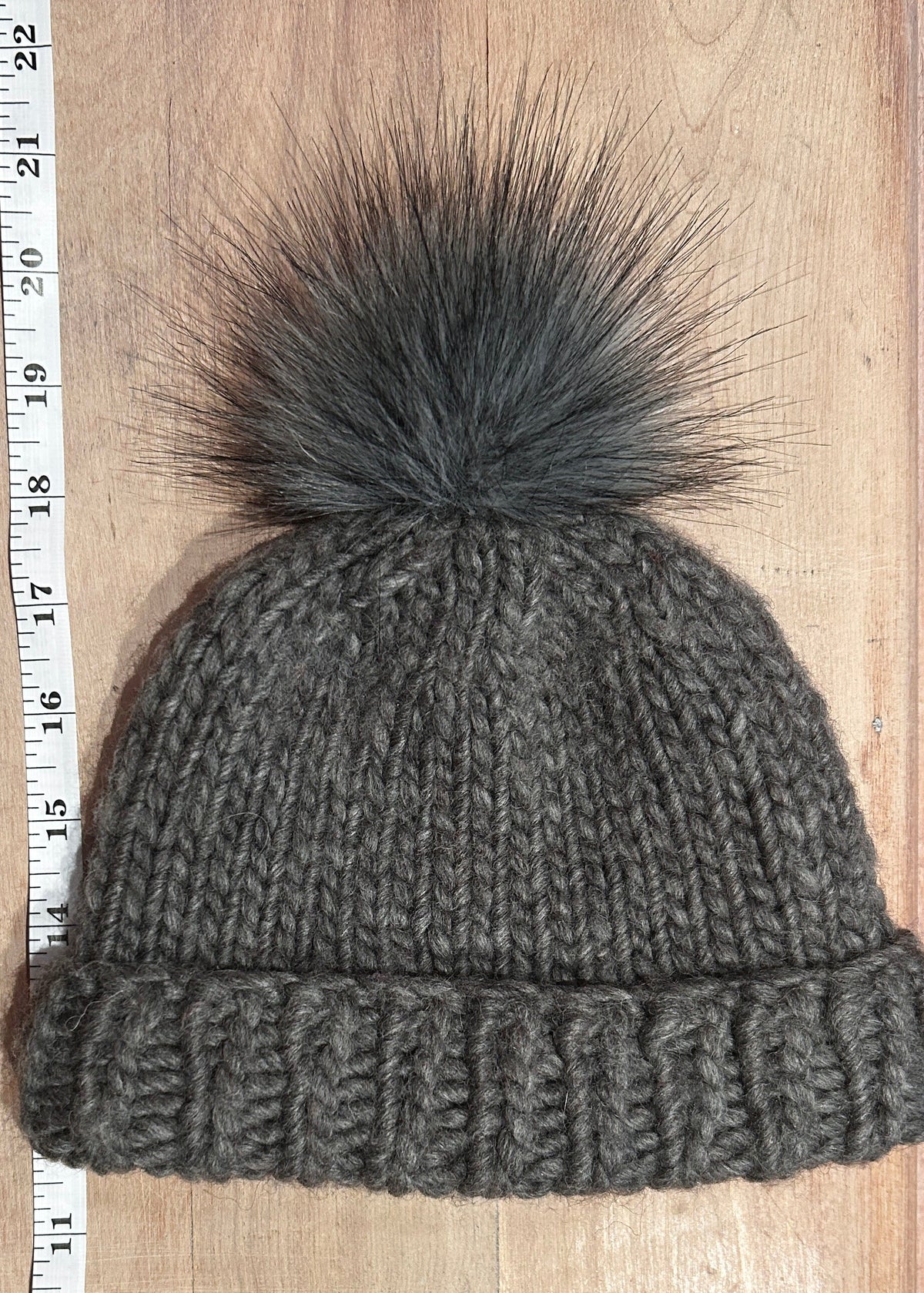 A small dark gray knit beanie with a small matching faux fur pom-pom, laid flat on a light wood surface next to a measuring tape ruler.