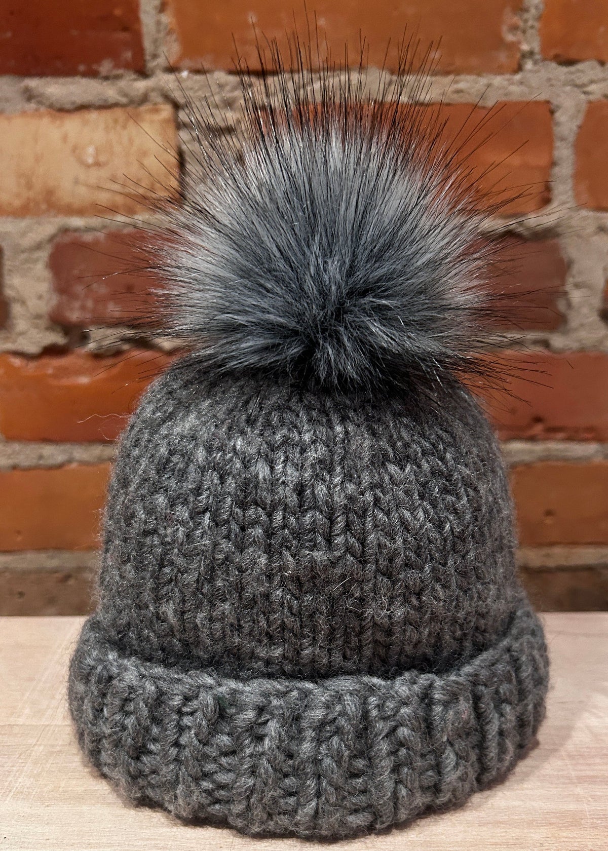 A small dark gray knit beanie with a small fluffy gray faux fur pom-pom, displayed on a light wood surface in front of a rustic red brick wall.