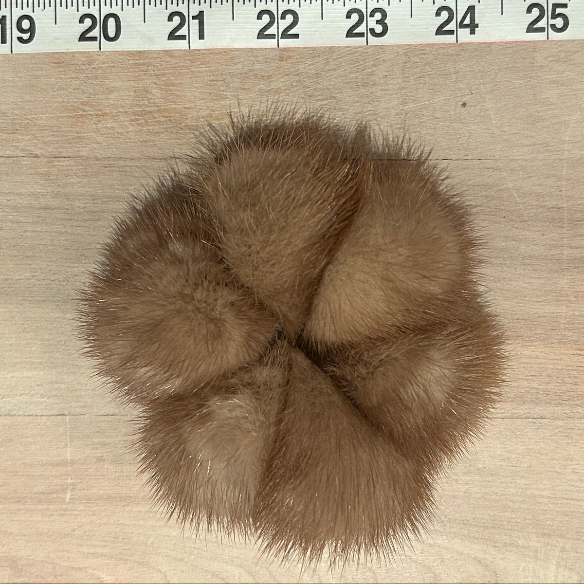A gold mink pom pom scrunchie hair accessory, photographed from above on a light wood surface with a measuring tape visible along the top edge for scale.