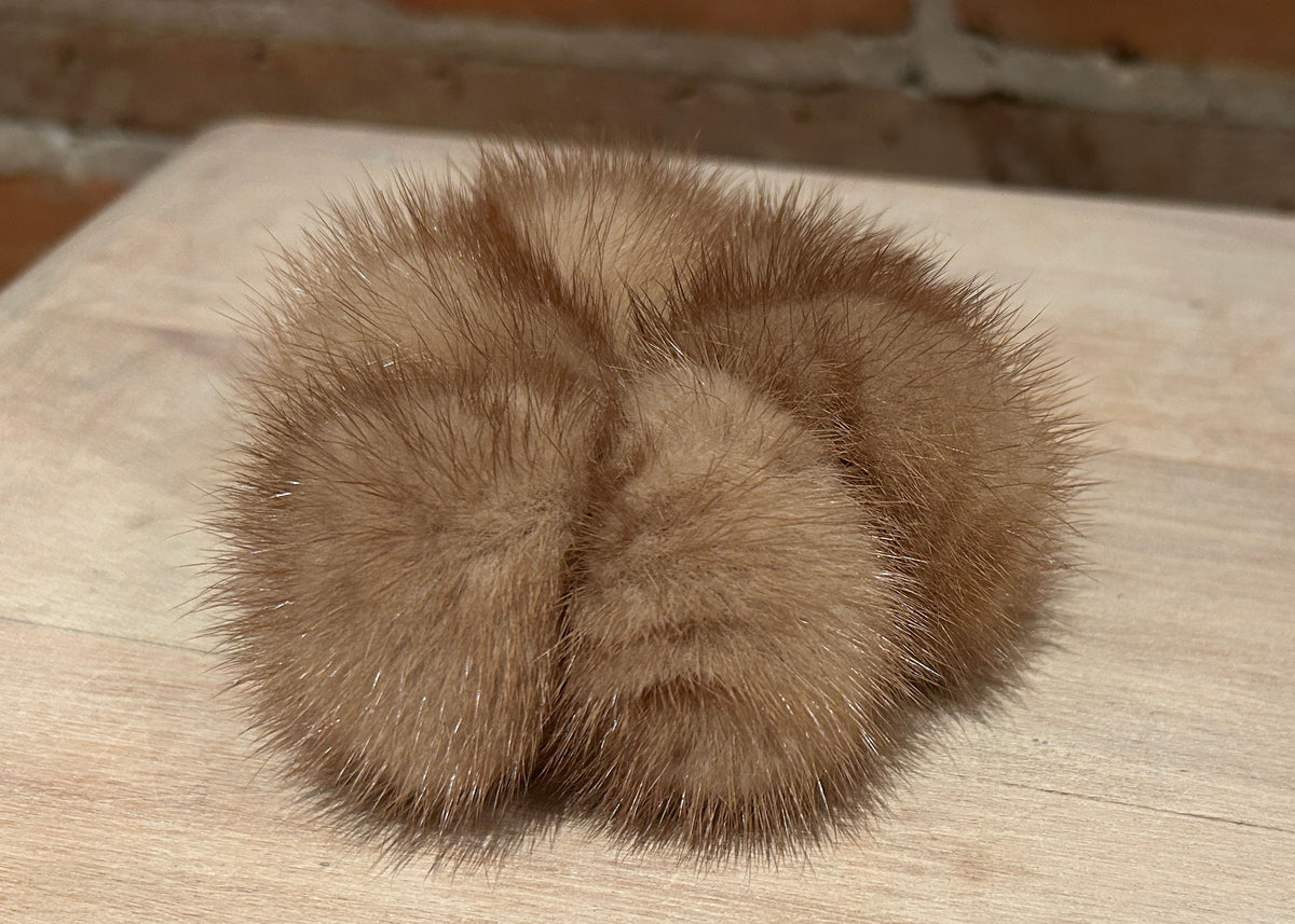 A gold mink scrunchie accessory resting on a light wood tabletop with a brick wall in the background.