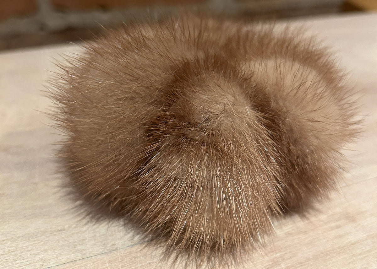 A fluffy gold mink fur pom-pom accessory resting on a light wood surface with a blurred brick wall background.