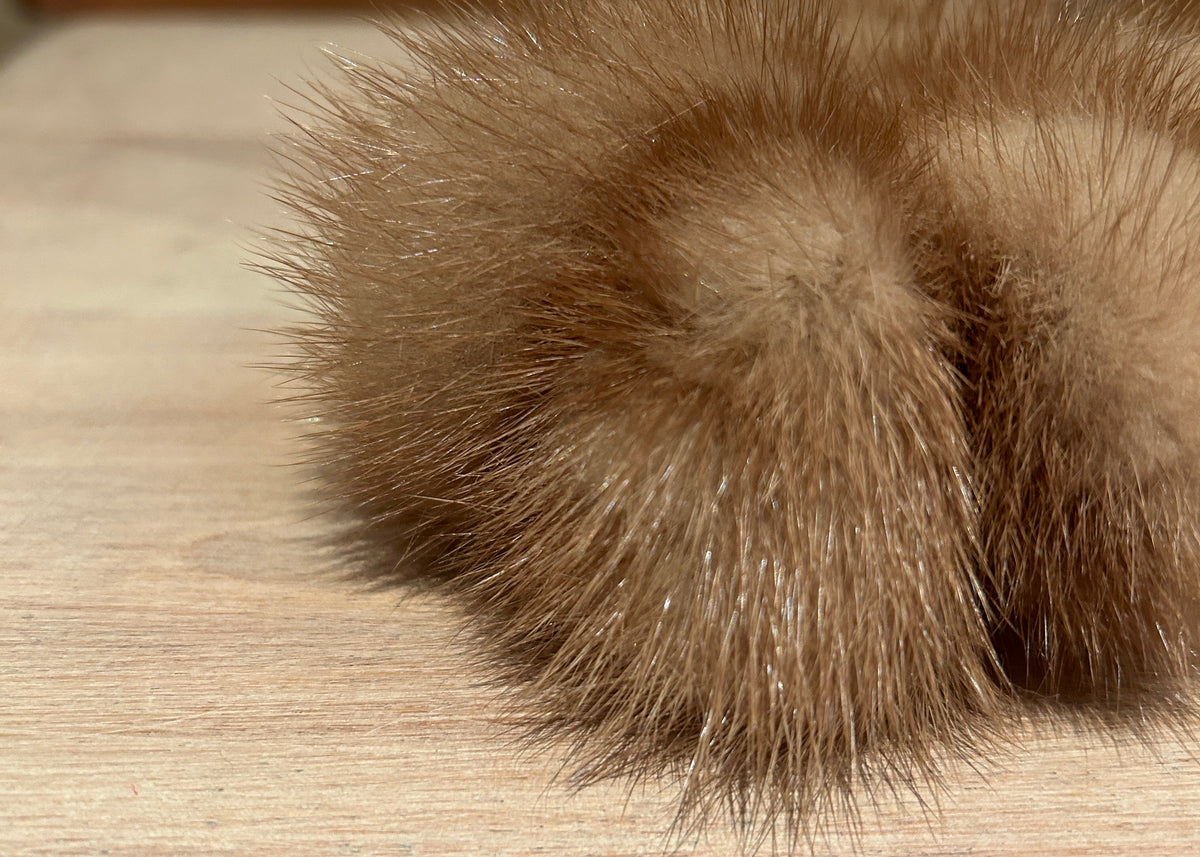 A close-up of a fluffy gold mink scrunchie resting on a light wood surface with a blurred brick wall background.