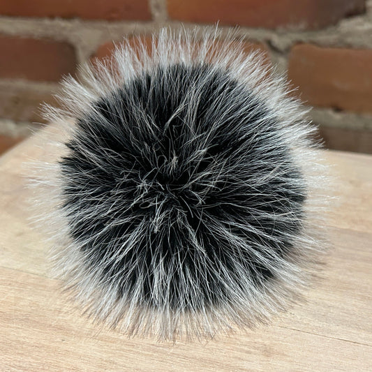 A round, fluffy pom-pom accessory made of black and silver-gray faux fur, resting on a light wood surface with a blurred brick wall background.