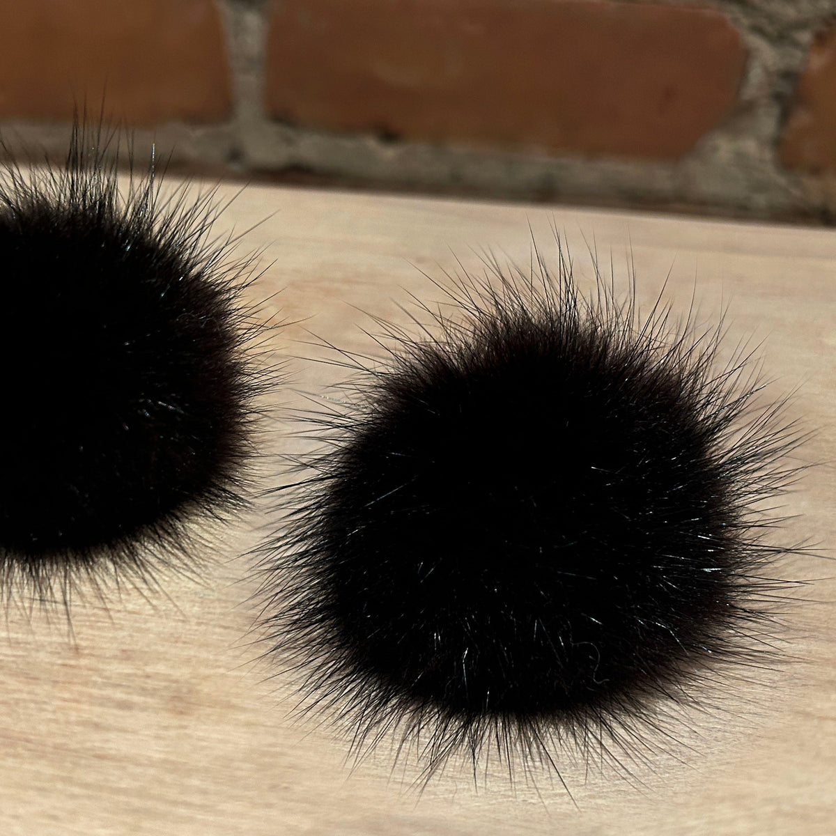 Two black mink fur pom-poms with long, spiky fur, placed on a light wood surface with a brick wall in the background.