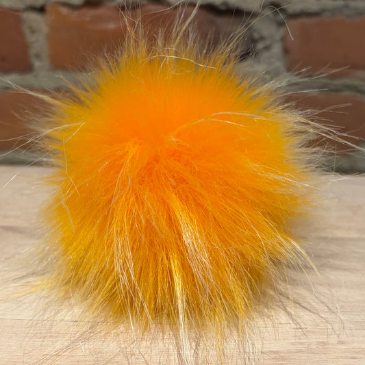 A bright orange fluffy faux-fur pom-pom ball resting on a light wood surface with a brick wall background.
