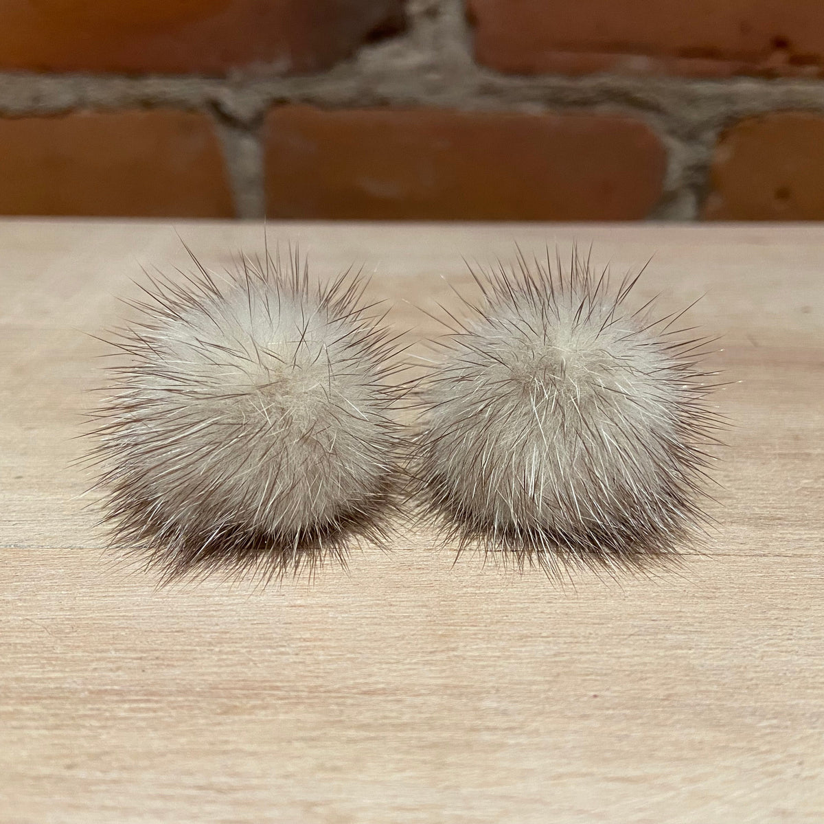 A pair of round, fluffy mink-fur pom-poms in a light beige and gray color, displayed on a light wood surface with a brick wall background.