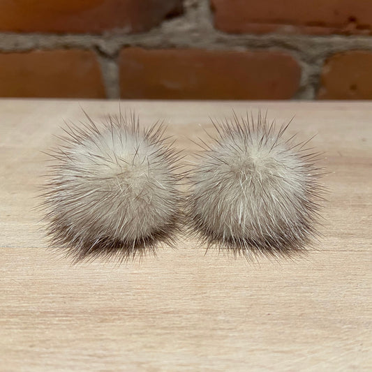 A pair of round, fluffy mink-fur pom-poms in a light beige and gray color, displayed on a light wood surface with a brick wall background.