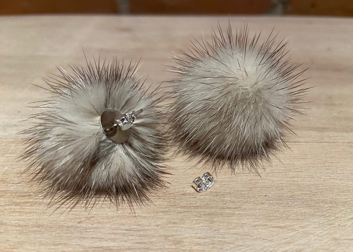 A pair of fluffy pom-pom earrings made from light gray and beige mink fur, with silver-tone metal backing and clear rhinestone studs, displayed on a light wood surface.