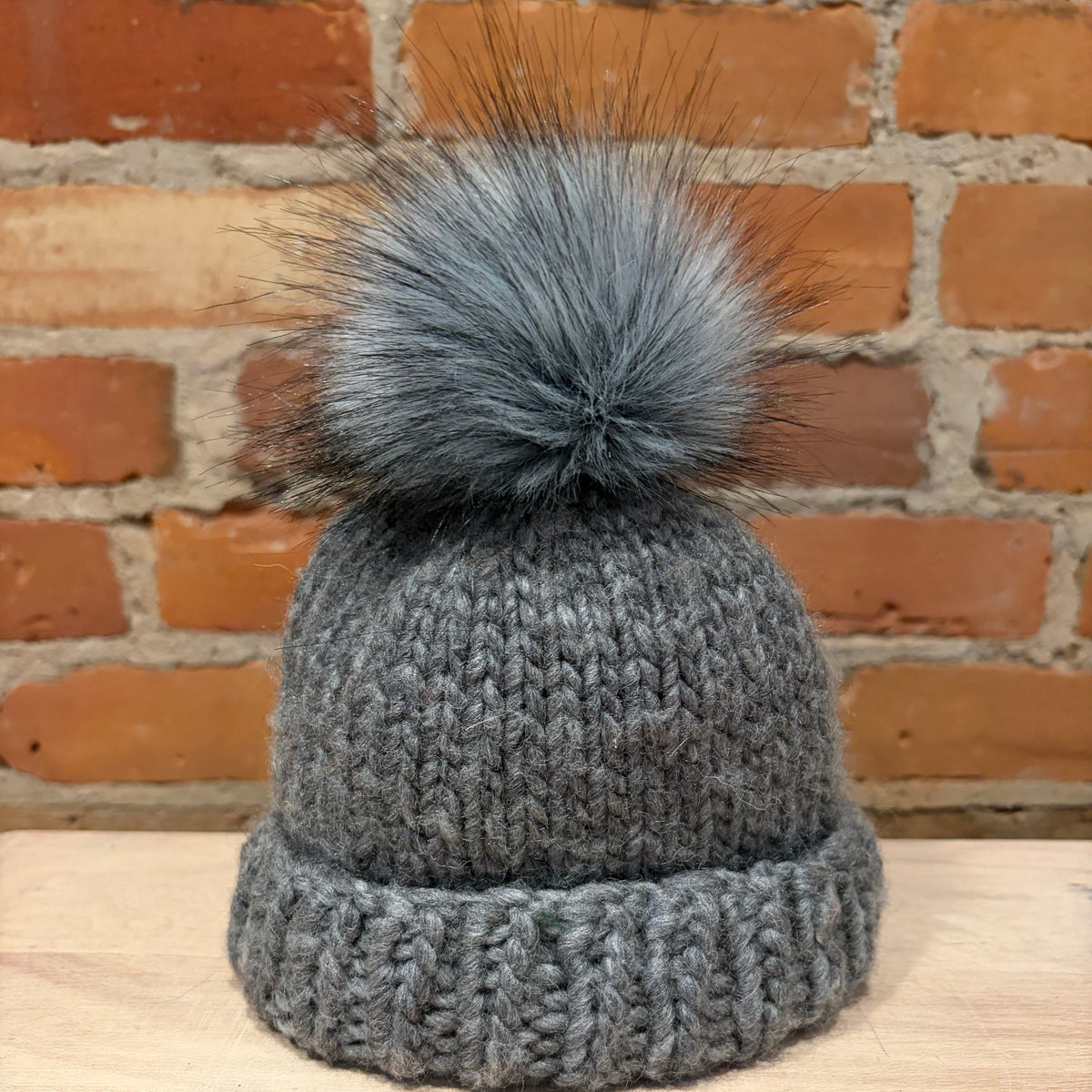A gray knit beanie hat with a large fluffy gray pom-pom, displayed on a light wood surface against a brick wall background.