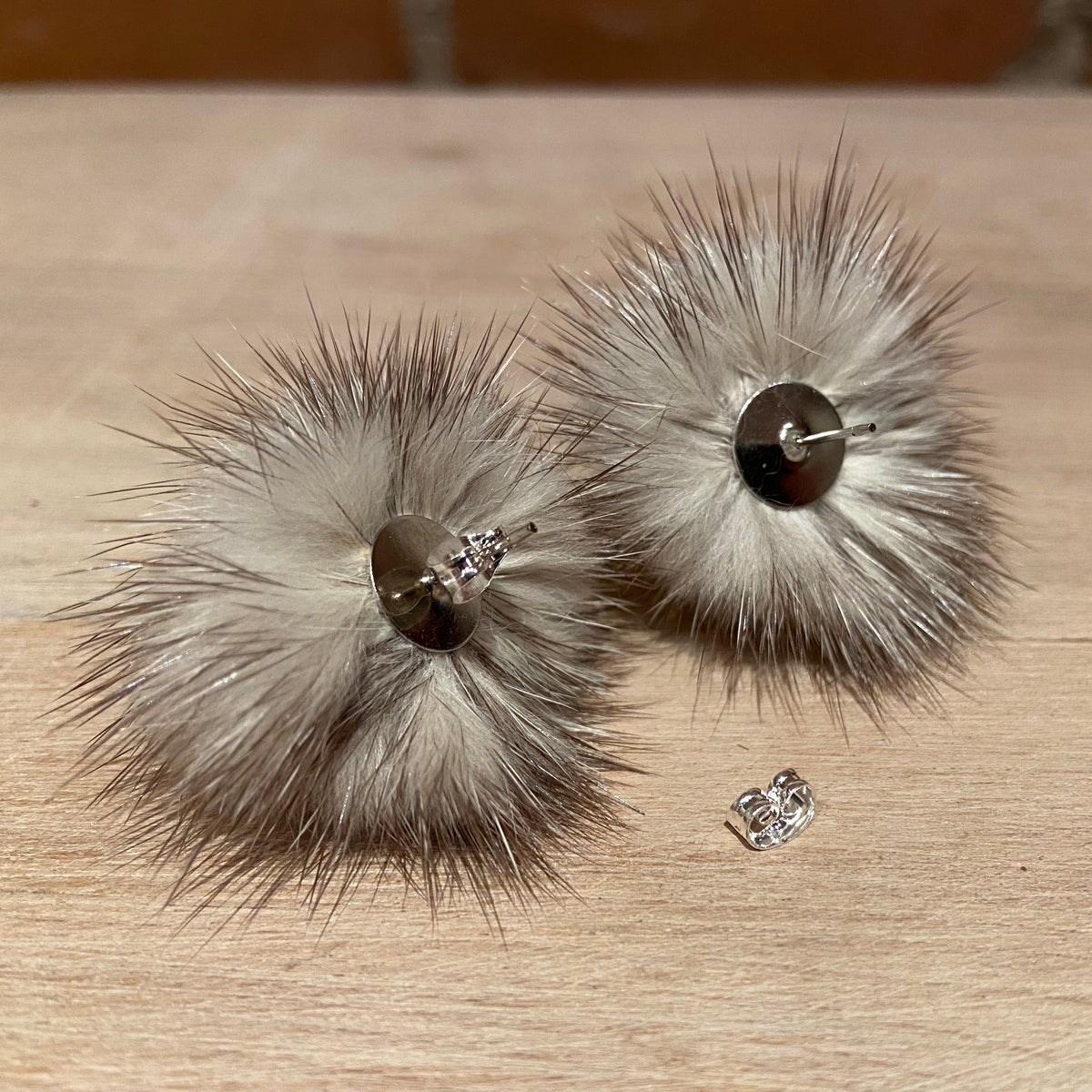 A pair of fluffy pom-pom earrings made from light brown and gray mink fur, with a dark brown button on the back and a small clear rhinestone stud placed in front, set on a light wood surface.