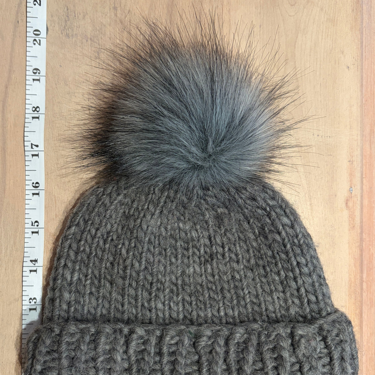 A gray ribbed knit beanie with a large fluffy gray faux-fur pom-pom, laid flat on a light wood surface next to a measuring tape.