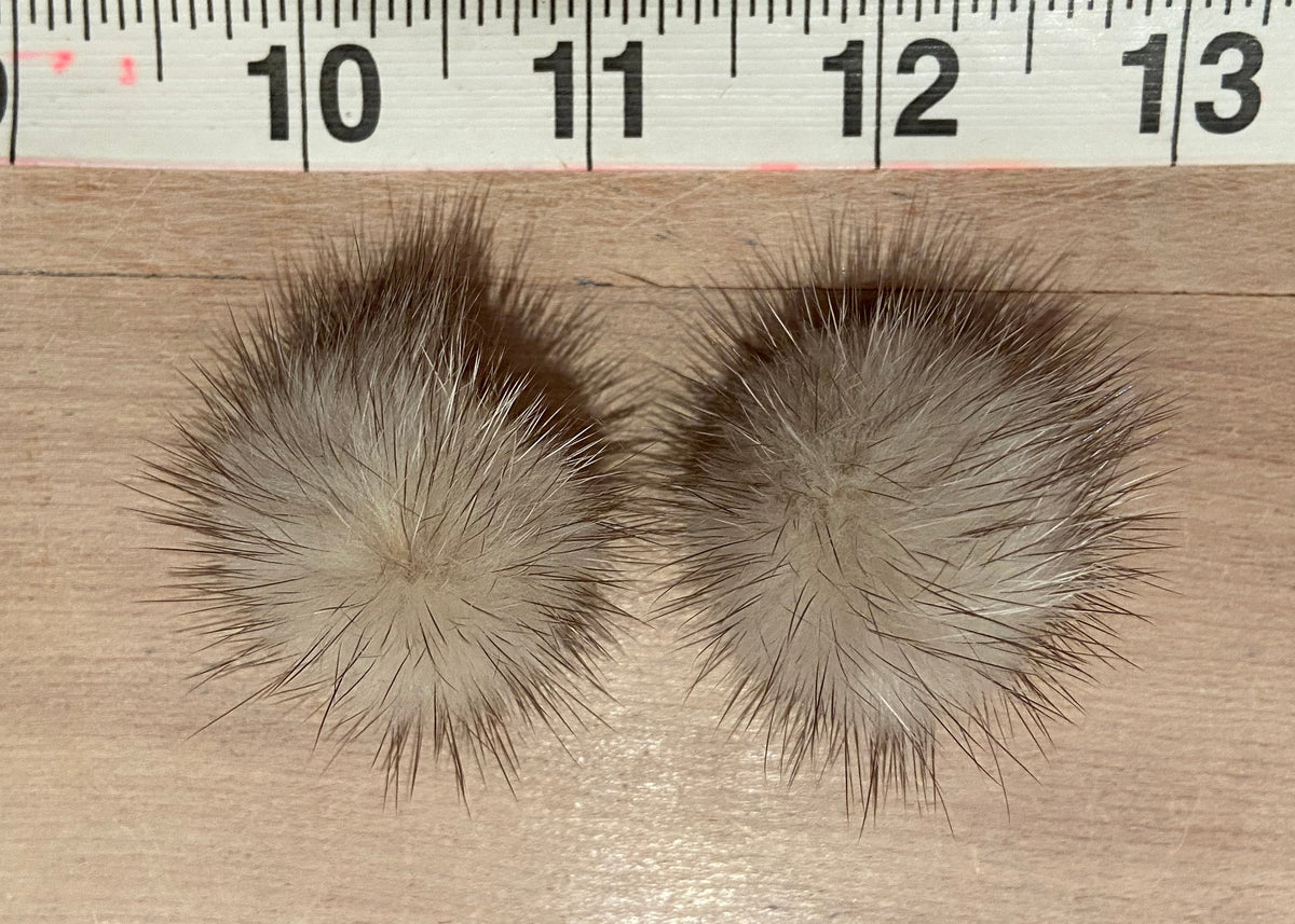 Two round fluffy mink-fur pom-poms in beige and brown, shown on a wooden surface with a measuring tape ruler at the top for scale.