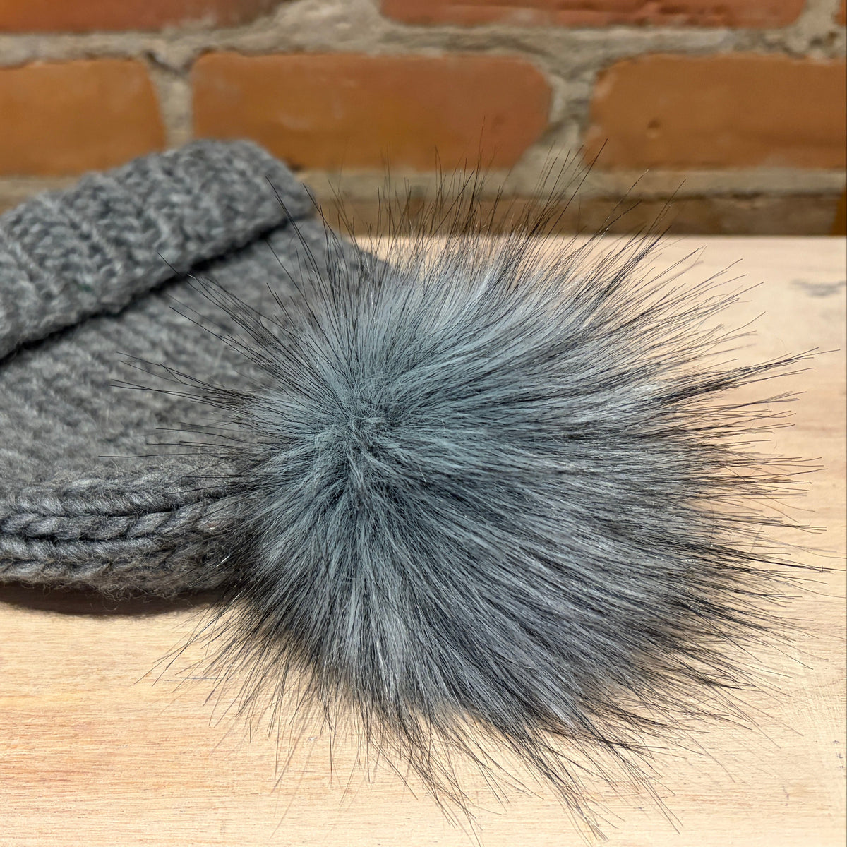 A gray knit beanie hat with a large fluffy gray faux-fur pom-pom, resting on a light wood surface with a brick wall background.