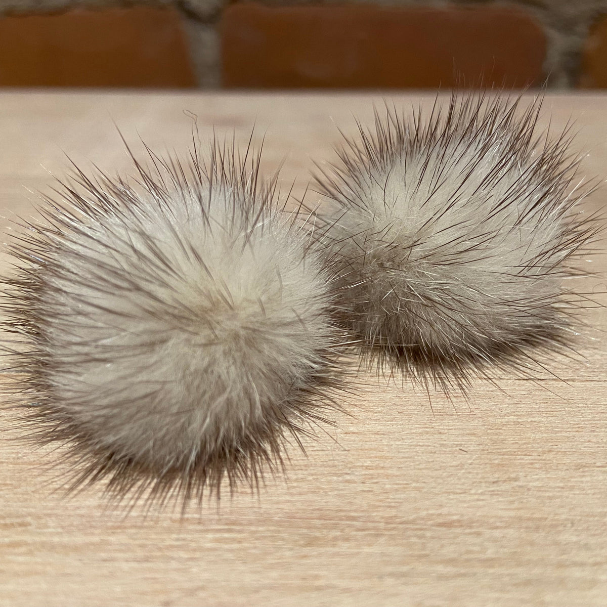 Two fluffy mink-fur pom-poms in a light beige and gray-brown color, resting on a light wood surface with a blurred brick wall background.