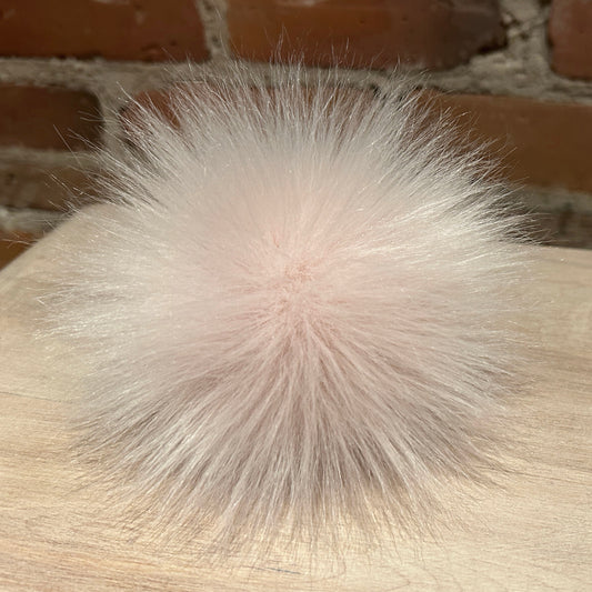 A fluffy faux-fur pom-pom in a pale blush pink color, resting on a light wood surface with a brick wall background.