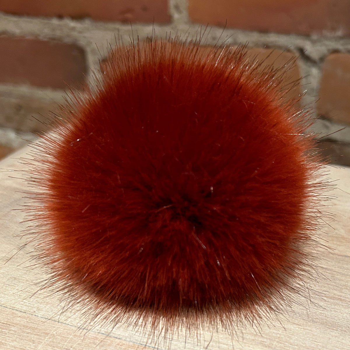 A round, fluffy pom-pom accessory in a rich rust-red or burnt orange color, resting on a light wood surface with a brick wall background.