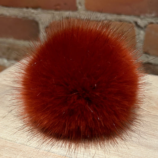 A round, fluffy pom-pom accessory in a rich rust-red or burnt orange color, resting on a light wood surface with a brick wall background.