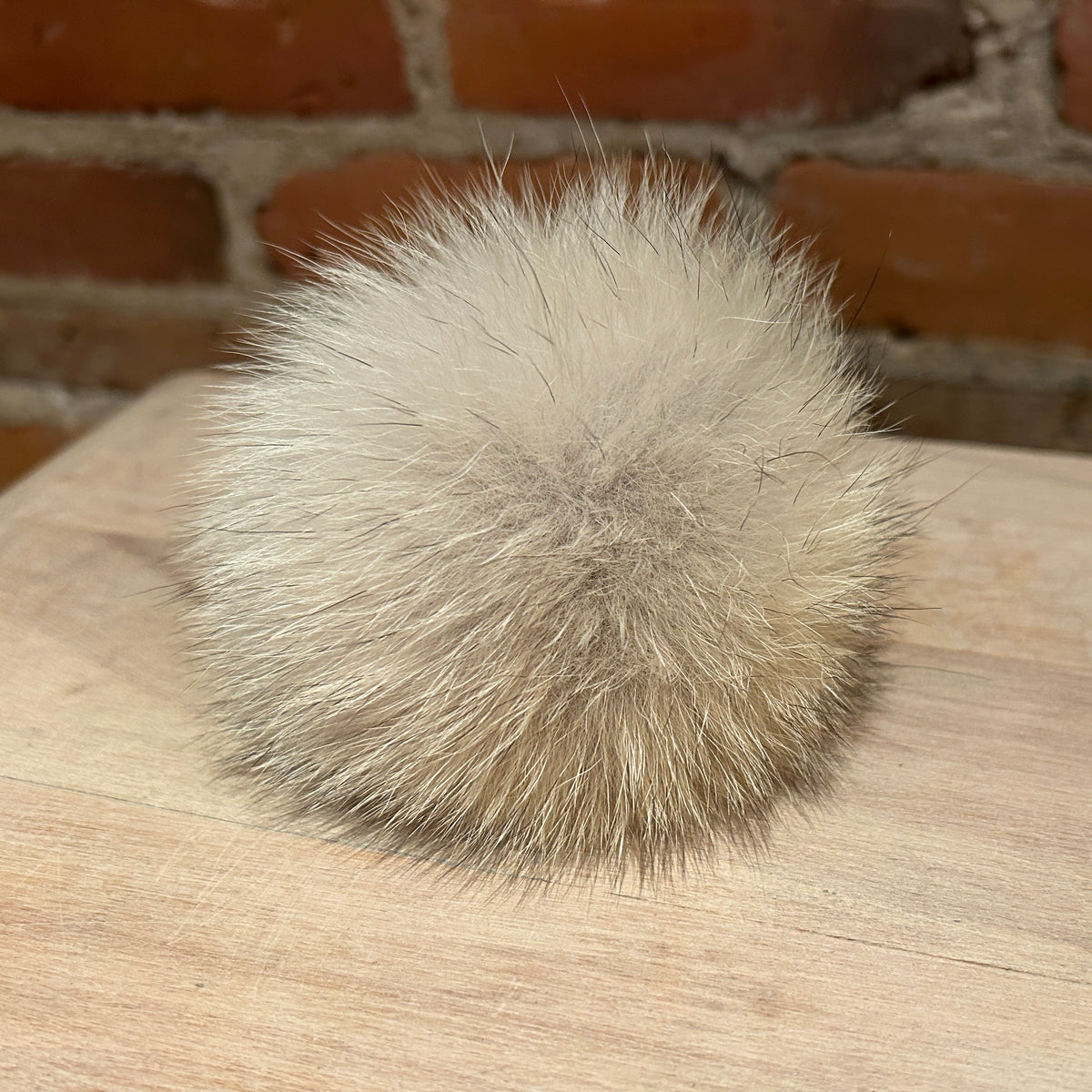 A fluffy coyote-fur pom-pom ball in a light beige and gray color, resting on a light wood surface with a blurred brick wall background.