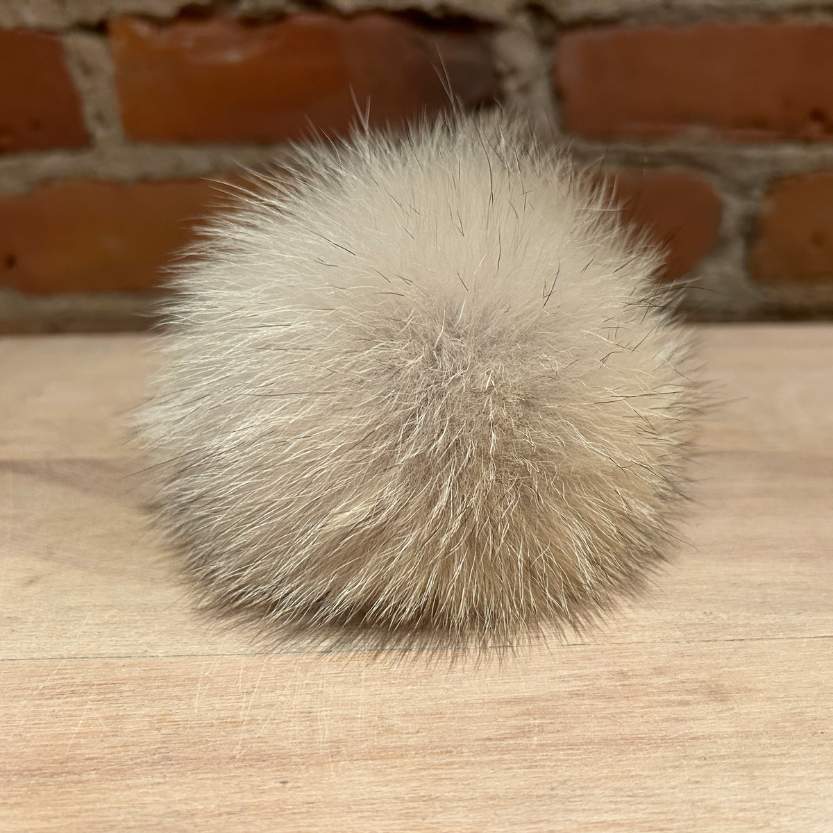 A fluffy pom-pom ball made of light beige coyote fur, resting on a light wood surface with a blurred brick wall background.