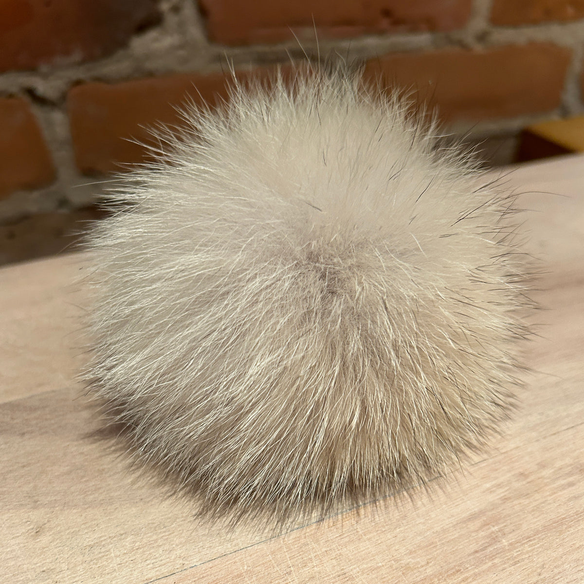 A fluffy pom-pom ball made of light beige and gray coyote fur, resting on a light wood surface with a blurred brick wall background.