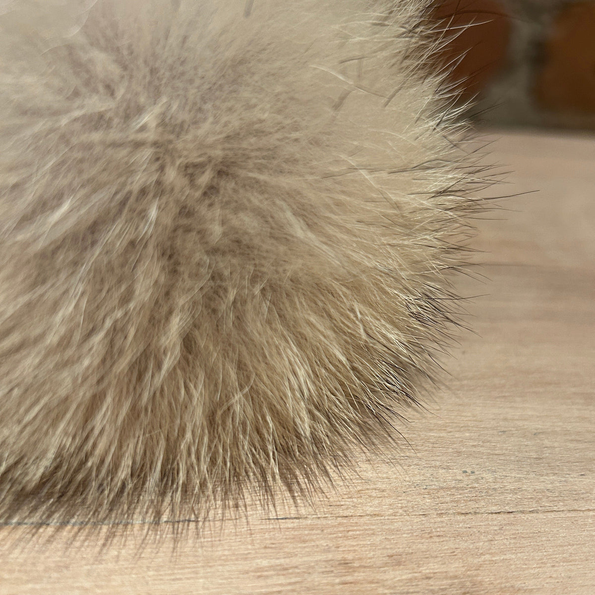 A close-up of a fluffy coyote fur pom-pom in a light beige and gray color, resting on a light wood surface.