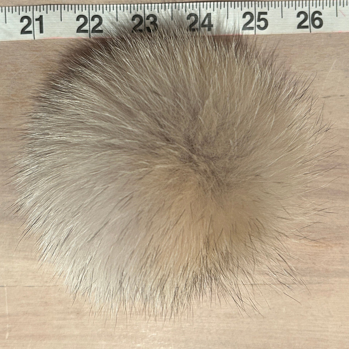 A round, fluffy coyote fur pom-pom in shades of beige, brown, and gray, shown on a light wood surface with a measuring ruler at the top for scale.