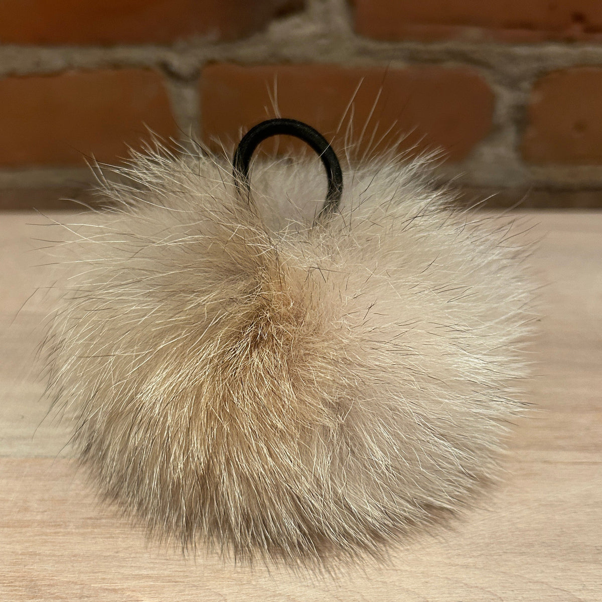 A fluffy coyote-fur pom-pom keychain in light beige and cream tones with a black metal key ring, resting on a light wood surface with a blurred brick wall background.