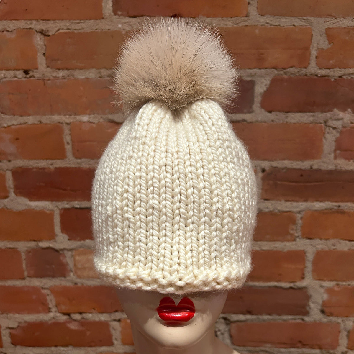 An off-white chunky knit beanie with a large beige coyote fur pom-pom, displayed on a mannequin head with red lips in front of a red brick wall.