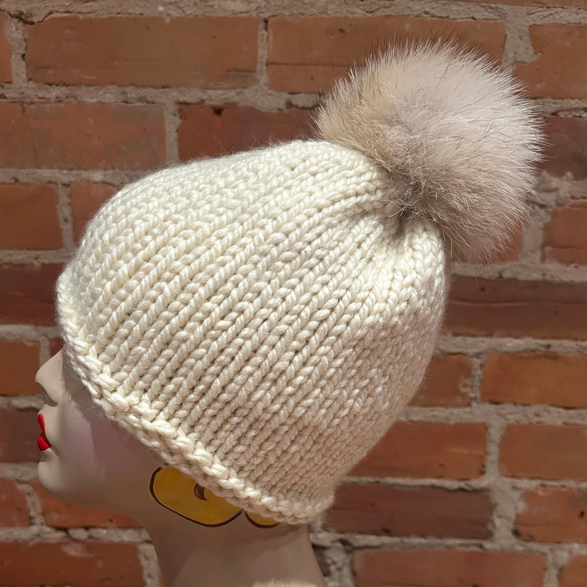 An off-white knit beanie with a large fluffy coyote fur pom-pom, displayed on a mannequin head against a red brick wall background.