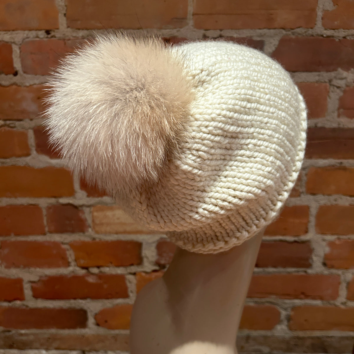 A cream-colored chunky knit beanie with a large fluffy coyote fur pom-pom, displayed on a mannequin head against a rustic red brick wall background.