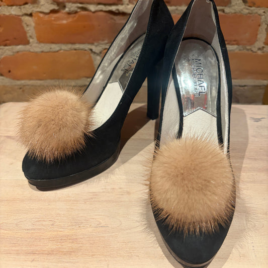 A pair of black high-heeled pumps with large beige mink-fur pom-poms on the toes, displayed on a wooden surface against a brick wall background.
