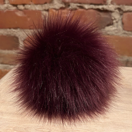 A fluffy pom-pom ball in deep burgundy faux fur, resting on a light wood surface with a brick wall background.