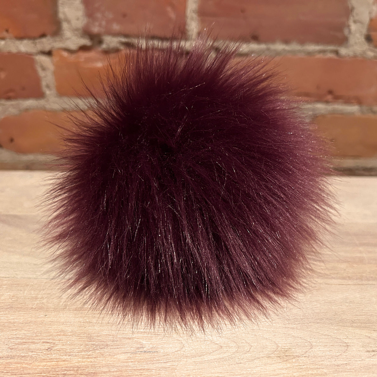 A dark burgundy faux fur pom-pom ball resting on a light wood surface with a blurred brick wall background.