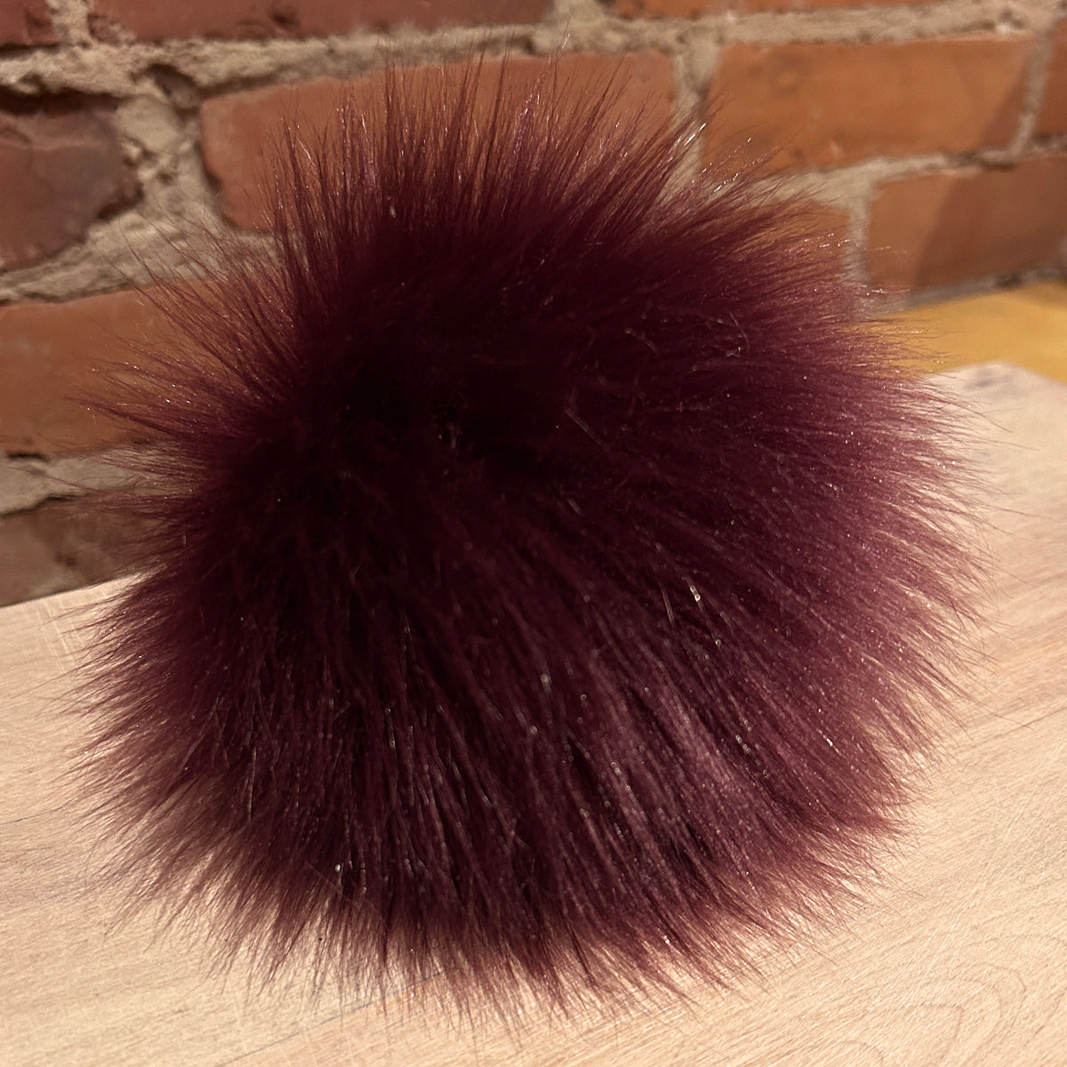 A large, fluffy pom-pom ball in a deep burgundy or maroon faux fur, resting on a light wood surface with a brick wall background.