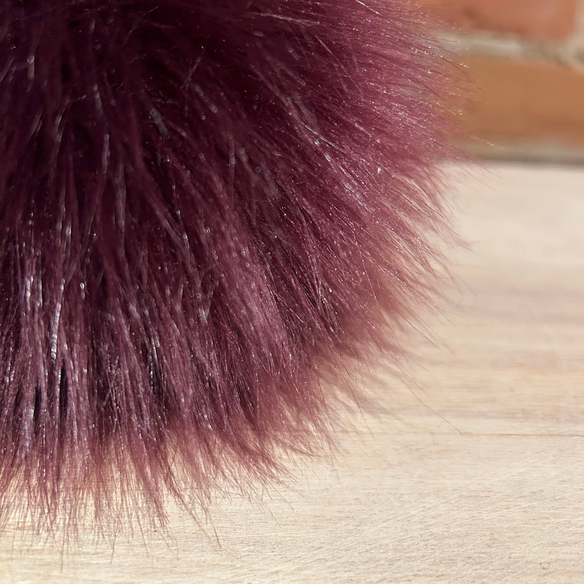 A close-up of a fluffy faux fur pom-pom in deep burgundy, resting on a light wood surface with a blurred brick wall background.