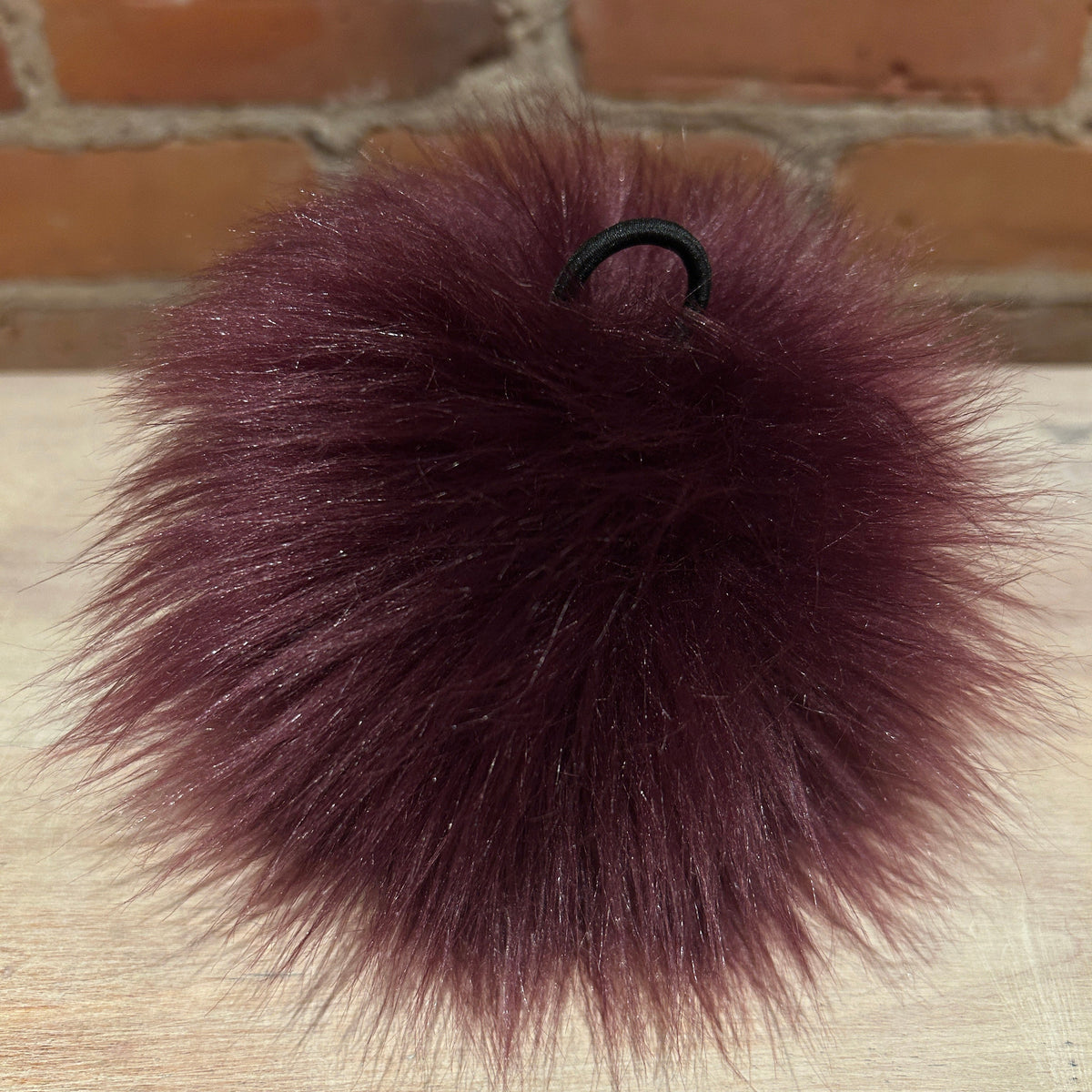 A fluffy pom-pom keychain in a deep burgundy color, featuring a black elastic loop at the top, resting on a light wood surface with a brick wall background.