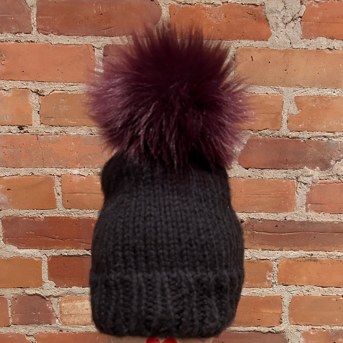 A dark charcoal gray knit beanie with a large, fluffy deep burgundy pom-pom, shown from the back against a rustic red brick wall.