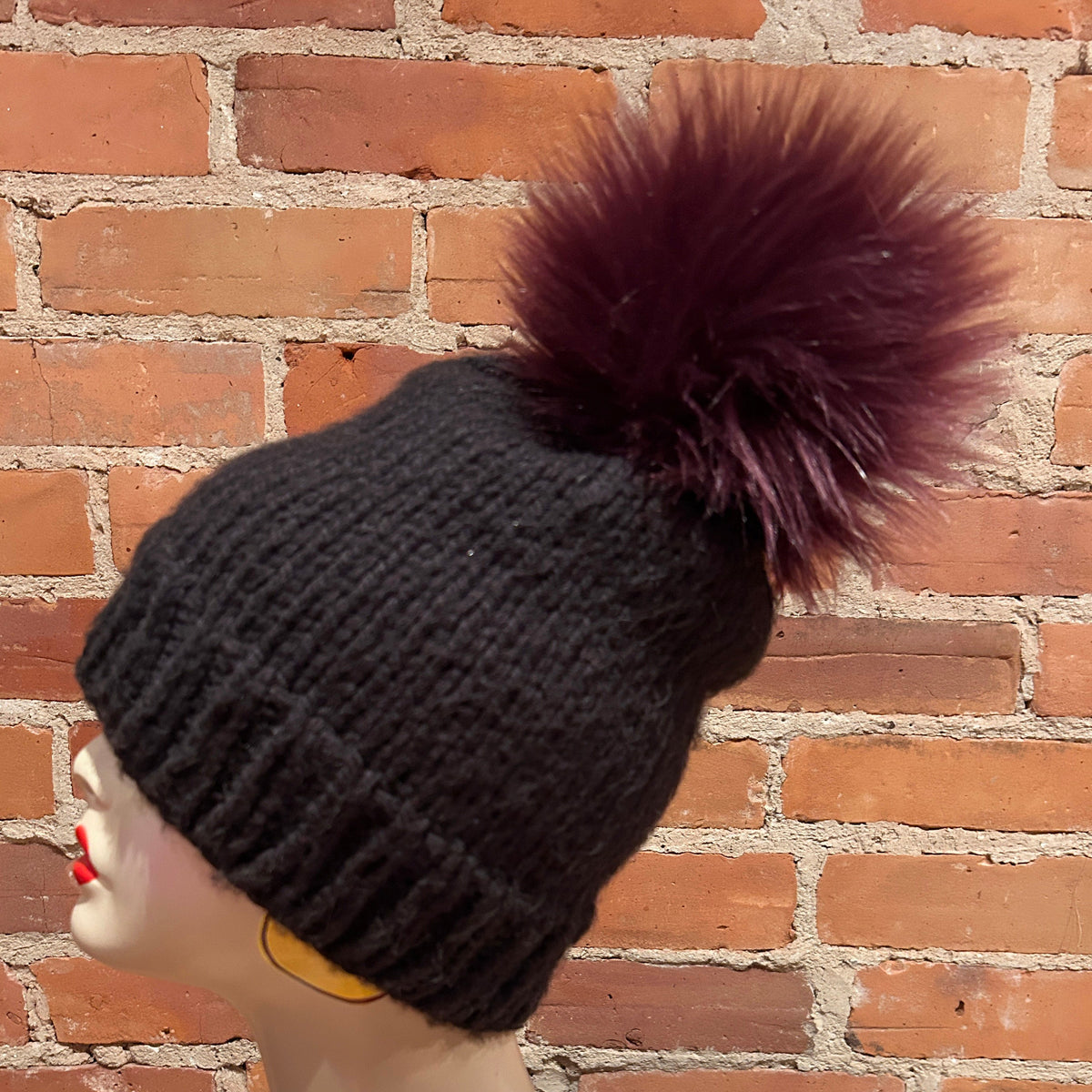 A black ribbed knit beanie with a large dark plum-burgundy faux fur pom-pom, displayed on a mannequin head against a red brick wall background.