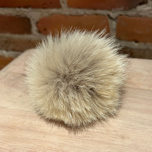 A fluffy beige coyote-fur pom-pom ball resting on a light wood surface with a brick wall in the background.