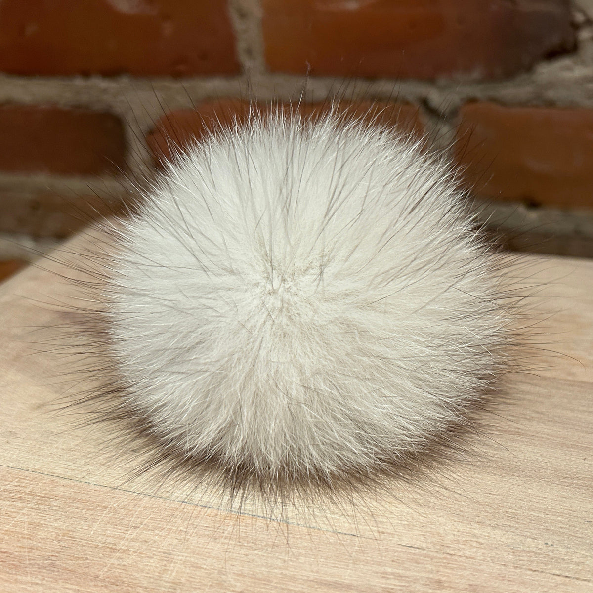 A fluffy white fox fur pom-pom ball resting on a light wood surface with a blurred brick wall background.