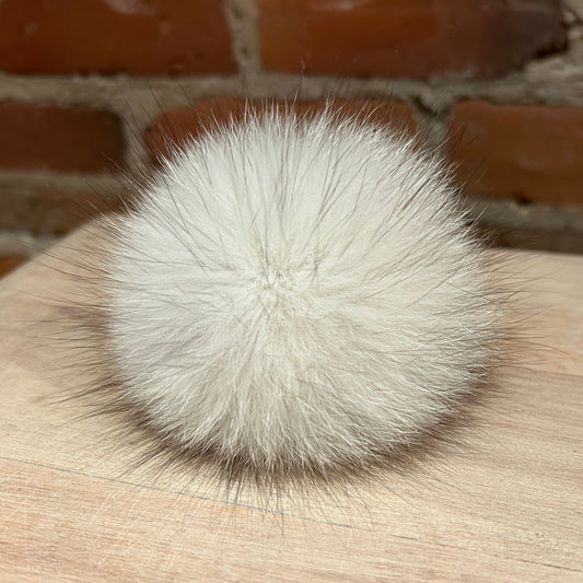 A fluffy white fox fur pom-pom ball resting on a light wood surface with a blurred brick wall background.