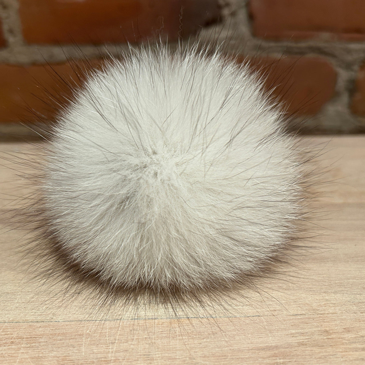 A fluffy white fox fur pom-pom ball with dark-tipped fibers, resting on a light wood surface with a brick wall background.