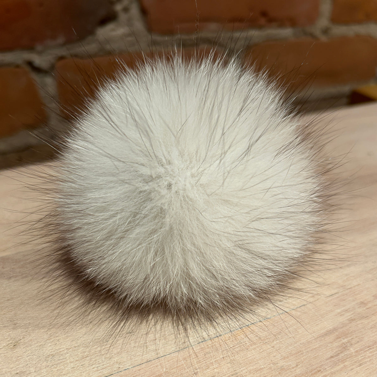 A fluffy white fox  fur pom-pom ball with long dark guard hairs, resting on a light wood surface with a brick wall background.