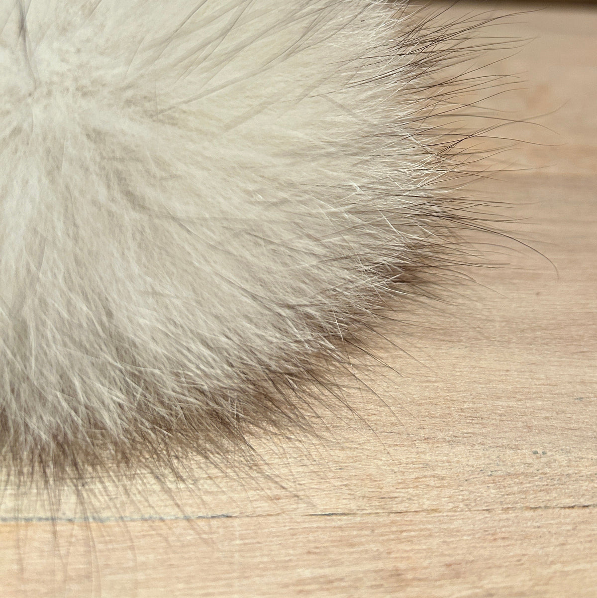 A close-up of a fluffy white fox fur pom-pom accessory resting on a light wood surface with a blurred brick wall background.