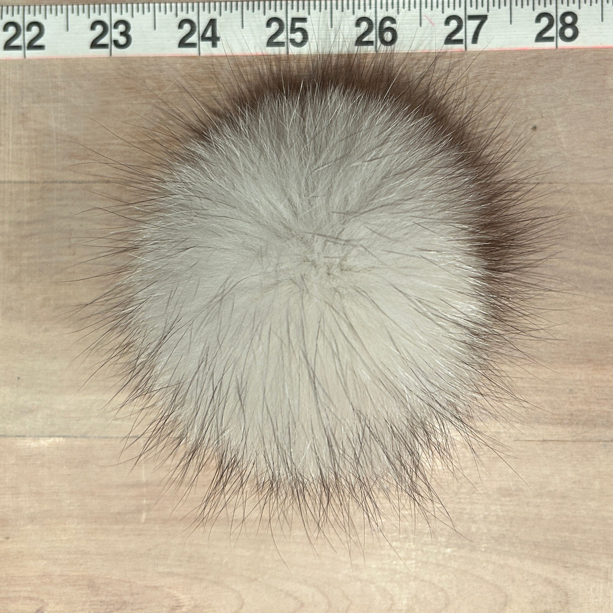 A round fluffy blue fox-fur pom-pom in light gray with dark brown tips, shown on a light wood surface with a measuring tape visible at the top for scale.