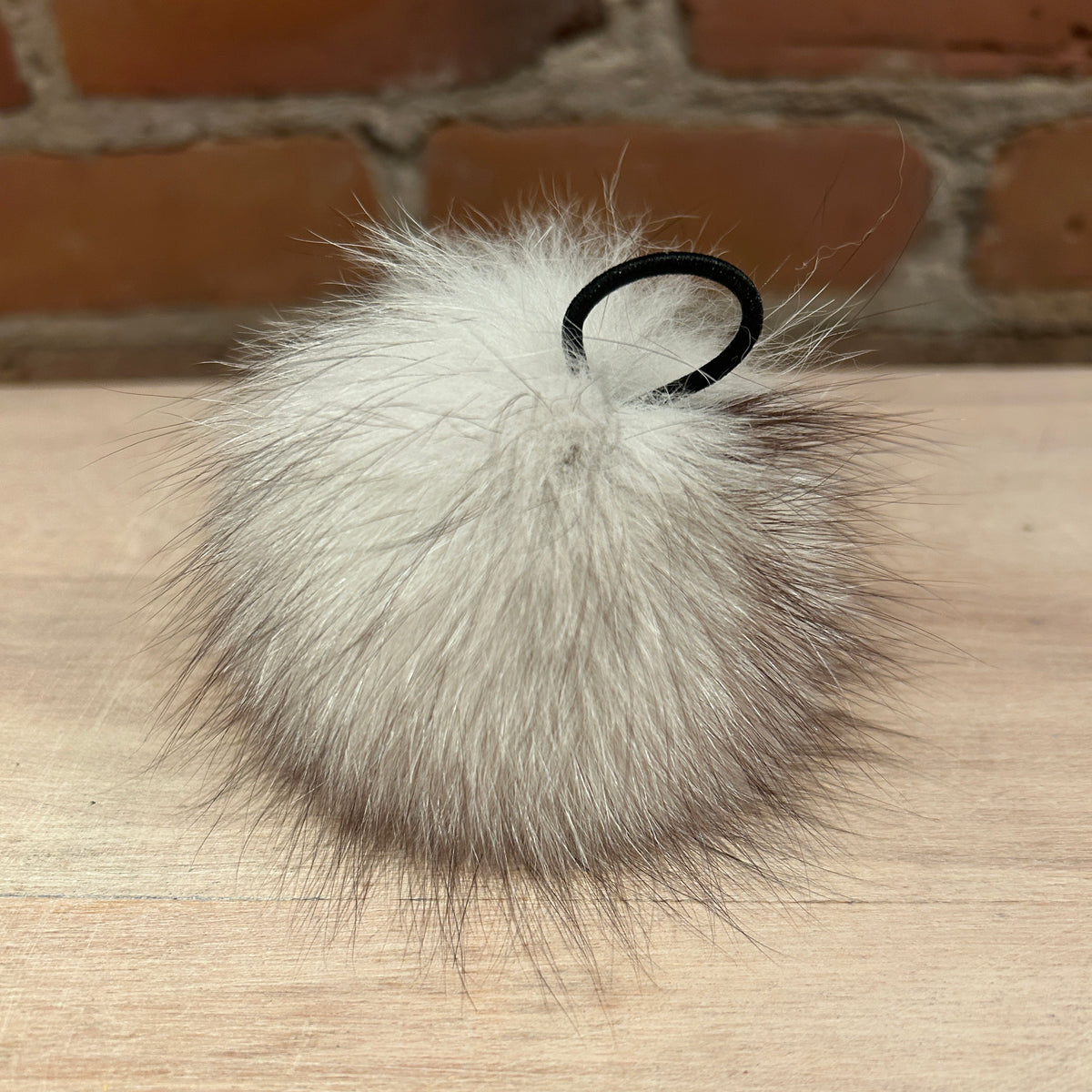 A fluffy blue fox-fur pom-pom , attached to a black elastic loop, resting on a light wood surface with a brick wall background.