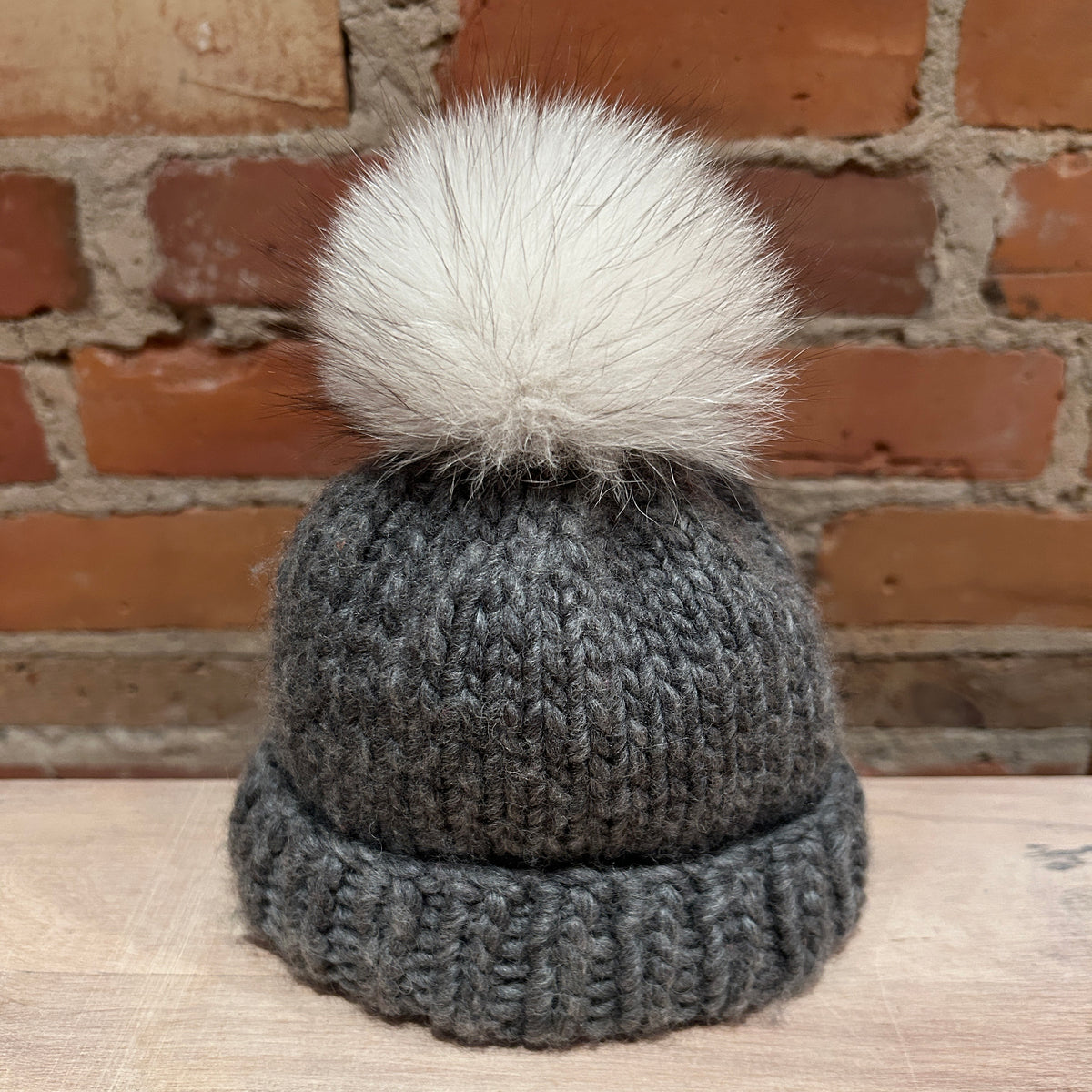A small dark gray knit beanie with a small white fox fur pom-pom, displayed on a light wooden surface in front of a brick wall background.