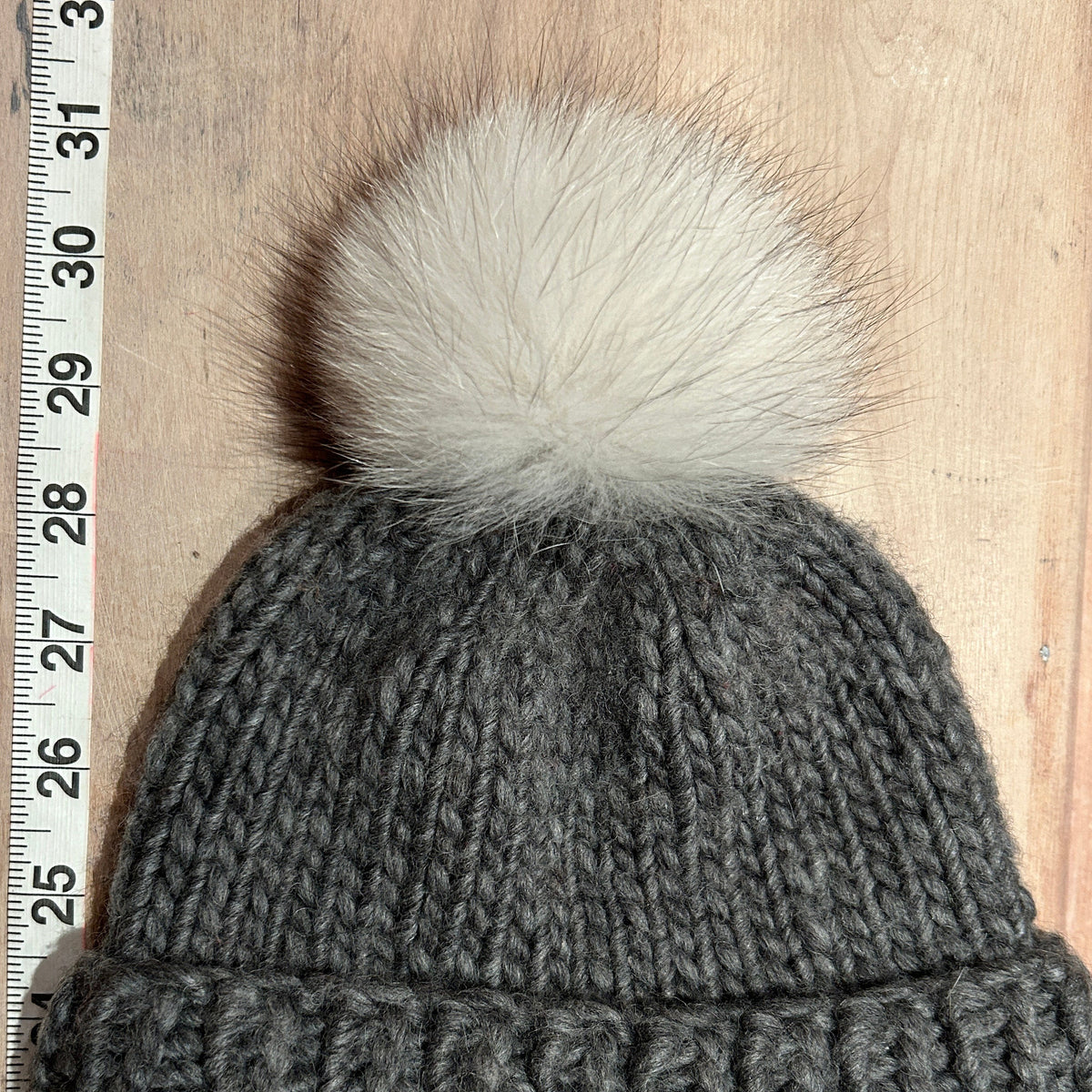 A small dark gray ribbed knit beanie with a small fluffy cream pom-pom, shown flat on a light wood surface next to a measuring ruler.